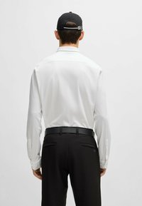 White long-sleeve shirt with a smooth texture, paired with black trousers and a black cap, seen from the back. Features a belt and two rear pockets.