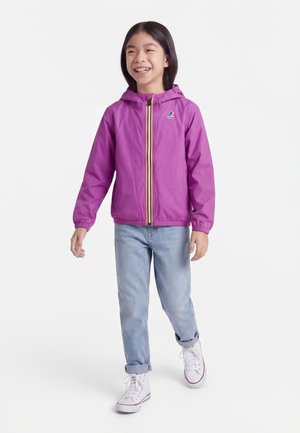 Mosolygó gyermek sétál, aki élénk lila kapucnis dzsekit, világoskék, hajtókás farmert és fehér magas szárú tornacipőt visel egyszínű háttér előtt.