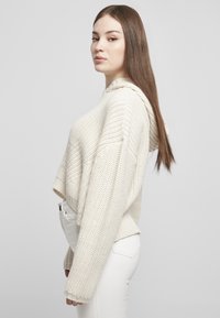 Kort hoodie i kremfarget strikkemateriale med teksturert overflate og hette. Har senkede skuldre og ribbestrikkede mansjetter. Kombinert med hvite bukser.