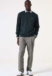 Sudadera verde oscuro con cuello redondo, con un logotipo en el pecho, superpuesta a una camisa blanca con estampado. Pantalones gris claro y zapatillas blancas.
