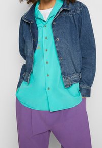 Denimjacka i mörkblått, med krage och knappar; turkos skjorta med knappar och en subtil design; lila byxor med avslappnad passform.