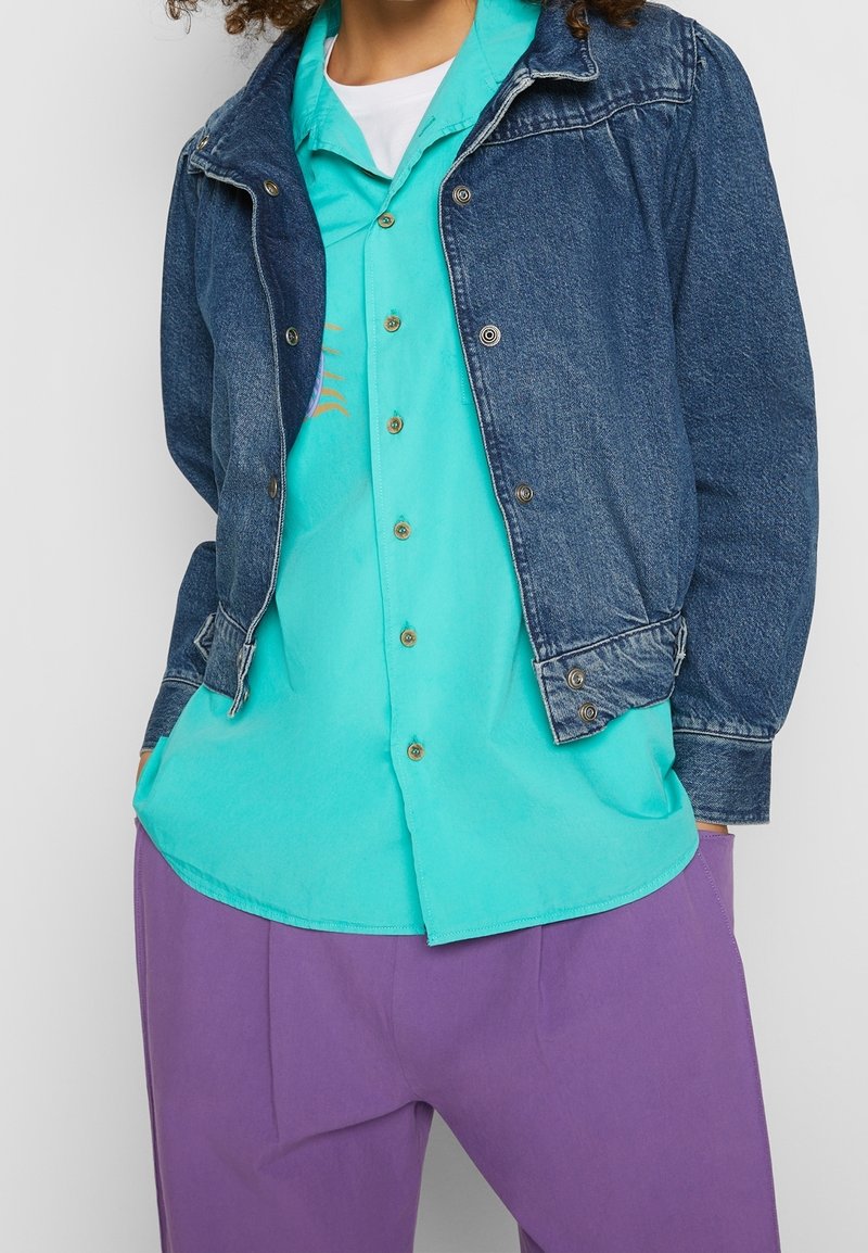 Denimjacka i mörkblått, med krage och knappar; turkos skjorta med knappar och en subtil design; lila byxor med avslappnad passform.