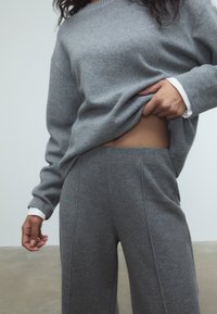 Sivý sveter s textúrovaným povrchom, s dlhými rukávmi a bielymi manžetami, skombinovaný s rovnakými sivými nohavicami s hladkým povrchom.