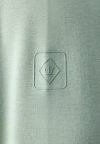 Tejido verde claro con una textura suave presenta un logo en relieve en forma de diamante con una corona en el centro. Diseño simple y minimalista.