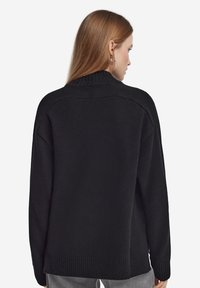 Svart stickad tröja med hög krage, sänkta axlar och ribbad nederkant. Mjuk textur med avslappnad passform, sedd bakifrån.