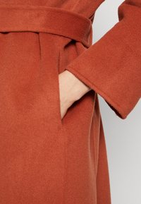 Mano che riposa all'interno della tasca di un cappotto di lana color ruggine con un ampio polsino e una cintura abbinata in vita.