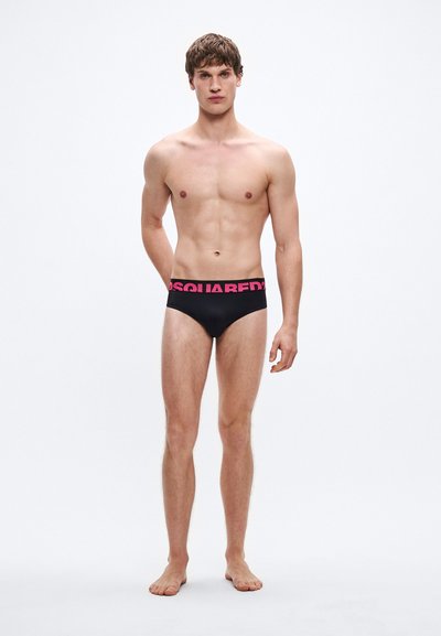 Meeste bikiinipüksid mustas värvitoonis, roosa logoga vööribaga, pehme kangas, keha järgnev lõige ja madal vöökohaga, esitatud meessoost modellil.