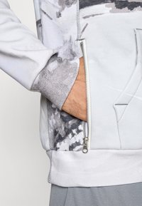 Lys grå zip-up sweatshirt med et tekstureret, mønstret område. Har ribbede manchetter og en kengurulomme. Lynlåsdetalje på siden.