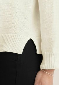 Neulotun kermanvärisen neuleen, jossa on ribbineulos, sivuhaara ja pitkät hihat. Yhdistetty kapeisiin mustiin housuihin ja hienostuneeseen rannekoruun.