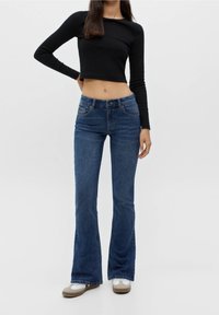 Top corto negro de manga larga combinado con jeans acampanados de un azul oscuro, con bolsillos y cierre de cremallera, complementado con zapatillas blancas y marrones.