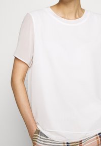 Top blanc à manches courtes avec un col rond. Tissu doux avec des manches transparentes. Associé à un pantalon à rayures dans des tons de beige, bleu et noir.