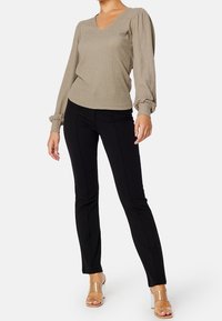 Donna che indossa un top lungo beige con texture e maniche a sbuffo, pantaloni neri slim e sandali trasparenti con tacco alto, in piedi contro uno sfondo bianco.