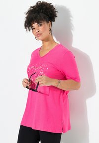 Camiseta de cuello en V rosa con mangas cortas, que presenta un diseño de corazones. La tela parece suave; el dobladillo tiene aberturas laterales. El modelo sostiene unas gafas de sol.
