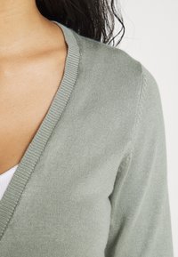 Sweter dzianinowy w kształcie litery V w delikatnym zielonym kolorze. Posiada prążkowane krawędzie oraz teksturowaną powierzchnię, podkreślającą gładkość i elastyczność materiału.