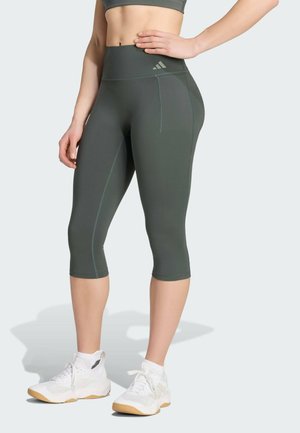 Person iført mørkegrønne højtaljede capri leggings og hvide træningssko, hånd hvilende på hoften, stående mod en ensfarvet baggrund.