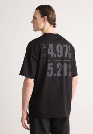 Zwarte katoenen T-shirt met grote grijze tekst op de achterkant: "Cash Price 4,97 9/10" en "Credit Price 5,28 9/10," met een losse pasvorm.