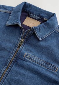 Chaqueta de mezclilla en azul oscuro, con cierre de cremallera, cuello puntiagudo, bolsillo en el pecho y detalles de costura en contraste. Textura suave.