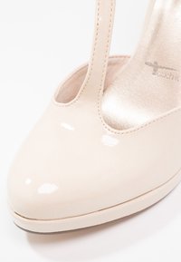 Beige lackläder högklackade sko med rund tå, utskärning på sidan och en slät, glansig yta. Dräneringsdetaljer längs kanterna.