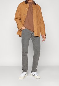 Béžová bavlněná bunda s hnědým límcem, hnědé tričko, šedé džíny a bílé tenisky s námořnickými pruhy. Neformální outfit předvedený na osobě.