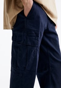 Marineblauwe cargobroek van zacht, textuurstof. Beschikt over meerdere zakken, waaronder een klepzak op de dij, en heeft een ontspannen pasvorm.