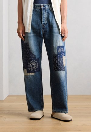 Persona che indossa jeans di denim blu con toppe di tessuto a motivo blu scuro su entrambe le gambe e scarpe beige slip-on, in piedi su pavimento in legno.