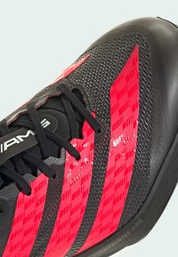 Sort atletiksko med røde, glansfulde mønsterdetaljer og sorte snørebånd. Udstyret med tekstureret materiale og mærket "LIGHTSTRIKE PRO" på sålen.