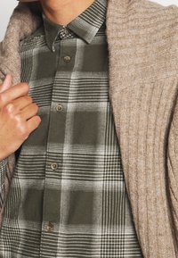 Beige Rippstrick-Cardigan, getragen über einem dunkelgrünen karierten Hemd mit weißen Akzenten. Das Hemd hat braune Knöpfe und markante Muster.