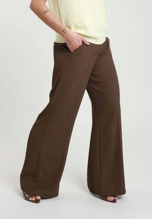 Persona che indossa pantaloni marroni a gamba larga e tacchi beige open toe, mano appoggiata nella tasca, top crema parzialmente visibile.