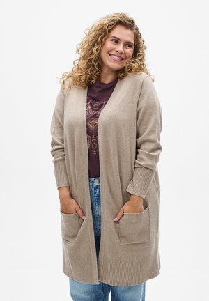 Long beige cardigan with ribbed cuffs, features two front pockets, made from a soft texture, paired with a dark purple graphic t-shirt and jeans.