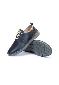 Scarpe in pelle blu navy con lacci bianchi, dettagli in marrone a contrasto e una suola in gomma grigia testurizzata con un motivo antiscivolo.