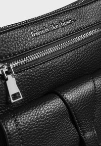 Primo piano di una borsa in pelle nera texturizzata con cerniera argentata e testo metallico che recita "amici come questi..." nella parte superiore.