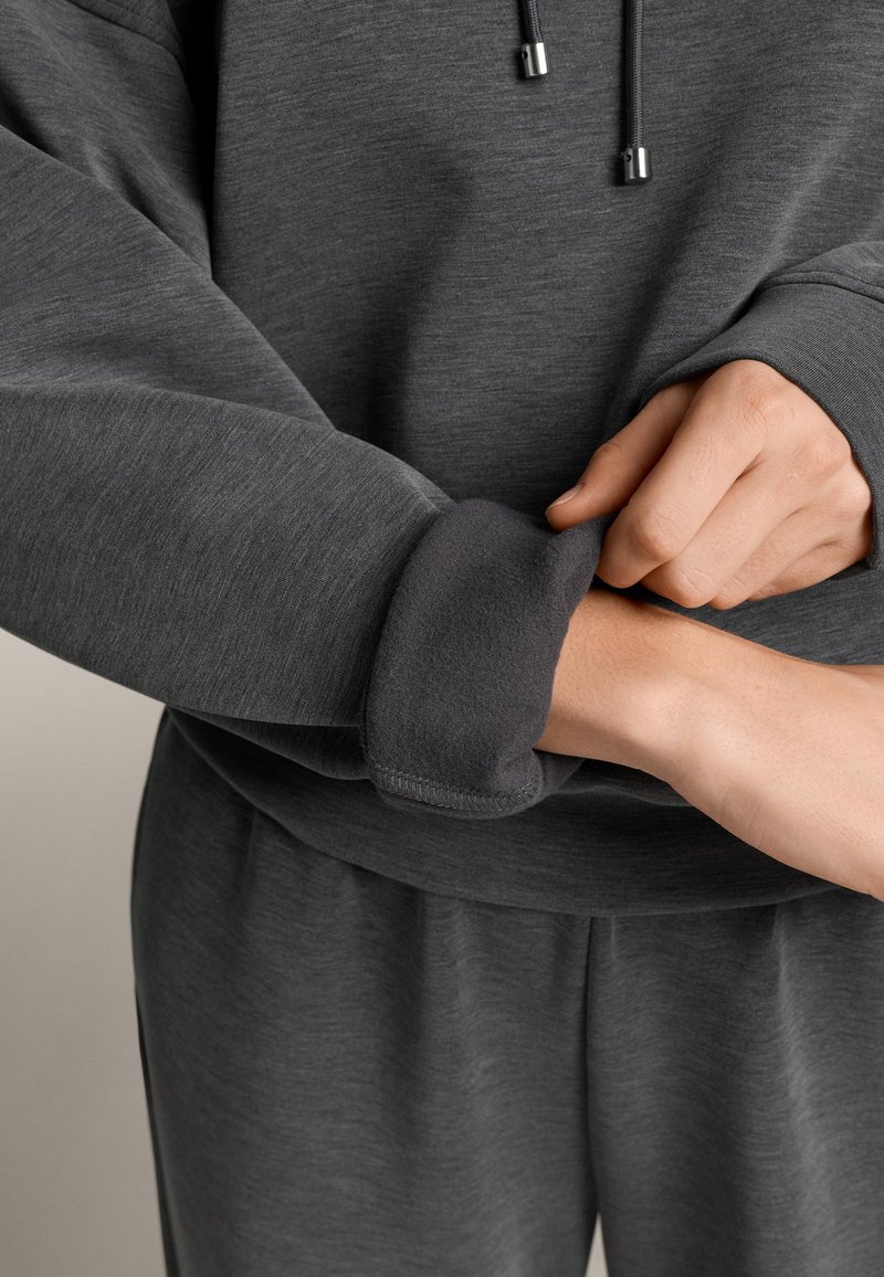 Hanorac gri închis cu textură netedă; are glugă, șnur și manșete moi. Tivul este ușor întors peste încheietură.