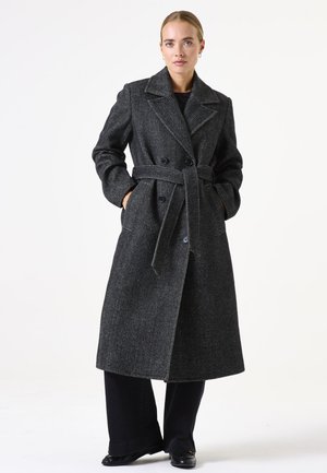 Manteau noir en chevrons à double boutonnage avec larges revers, une ceinture et des poches latérales, associé à un pantalon noir et des chaussures.