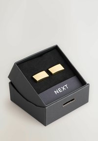 Guldfarvede rektangulære manchetknapper præsenteret i en sort æske med en blød indvendig foret, som har brandnavnet "NEXT" på låget.