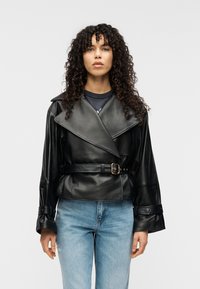 Μαύρο cropped δερμάτινο μπουφάν με μεγάλους γιακάδες, ζώνη στη μέση και φαρδιά μανίκια. Φορεμένο πάνω από σκοτεινό πουκάμισο, σε συνδυασμό με ανοιχτό μπλε τζιν.