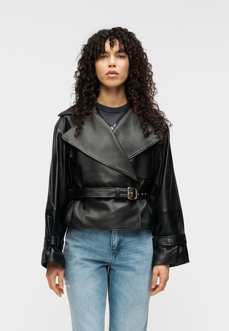 Μαύρο cropped δερμάτινο μπουφάν με μεγάλους γιακάδες, ζώνη στη μέση και φαρδιά μανίκια. Φορεμένο πάνω από σκοτεινό πουκάμισο, σε συνδυασμό με ανοιχτό μπλε τζιν.