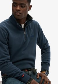 Granatowy polar z suwakiem w połowie długości, z haftowanym logo, nałożony na zieloną koszulę w kratkę, z zegarkiem na nadgarstku i dżinsami.