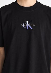 Crna pamučna majica s istaknutim ljubičastim i bijelim logotipom "Calvin Klein Jeans" i stiliziranim dizajnom "CK" na prsima.