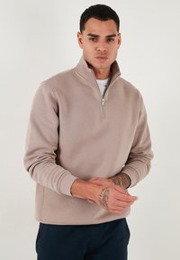 Sudadera con cremallera color marrón claro, con cuello alto, puños acanalados y una textura suave. Presenta un pequeño detalle de cremallera en el cuello.