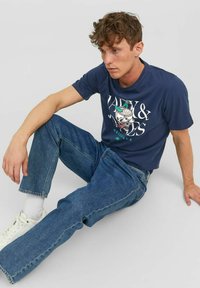 Camiseta de algodón azul marino con estampado gráfico, combinada con jeans de mezclilla azul claro. Modelo sentado contra un fondo neutro. Zapatillas blancas visibles.