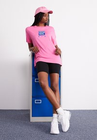 Růžové tričko Nike s modrým logem, černé cyklistické šortky, bílé tenisky a odpovídající růžová kšiltovka, na pozadí modré skříně s šuplíky.