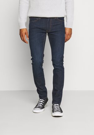 Man draagt donkerblauwe slim-fit jeans, zwart-witte hoge sneakers en een lichtgrijze geribbelde trui en staat op een grijze vloer.
