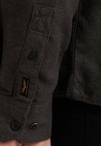 Donkergrijze flanellen shirtmanchet met een knoopsluiting, decoratieve stiksels en een gemarkeerd zwart label met gouden letters.