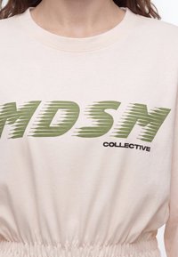 Gros plan d'une personne portant un haut beige avec un texte vert "MDSM" et noir "COLLECTIVE" imprimé sur la poitrine.