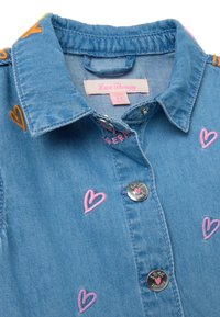 Denimhemd mit einem hellblauen Finish, verziert mit bestickten rosa und orangen Herzen, silbernen Druckknöpfen und einem Etikett mit der Aufschrift "Love Therapy".