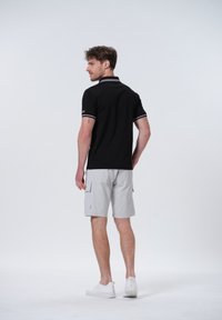 Camisa polo negra con ribete blanco, combinada con pantalones cortos de cargo gris claro. El modelo lleva zapatillas blancas. El fondo es liso y claro.