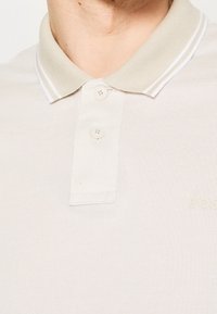 Gros plan d'une personne portant un polo beige avec un col et deux boutons, montrant une partie du cou et de la poitrine.