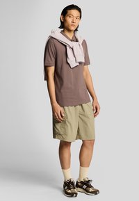 Camisa polo marrón, sudadera gris claro caída sobre los hombros, pantalones cortos beiges, calcetines crema y zapatillas marrones con detalles oscuros.