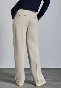 Beige Hochformat-Hosen mit lockerem Schnitt. Mit zwei Gesäßtaschen, glatter Textur und dezenten Faltendetails an jedem Bein.