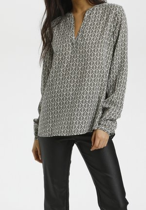 Woman wearing a black and white patterned long sleeve blouse paired with black leather pants, standing against a plain background.